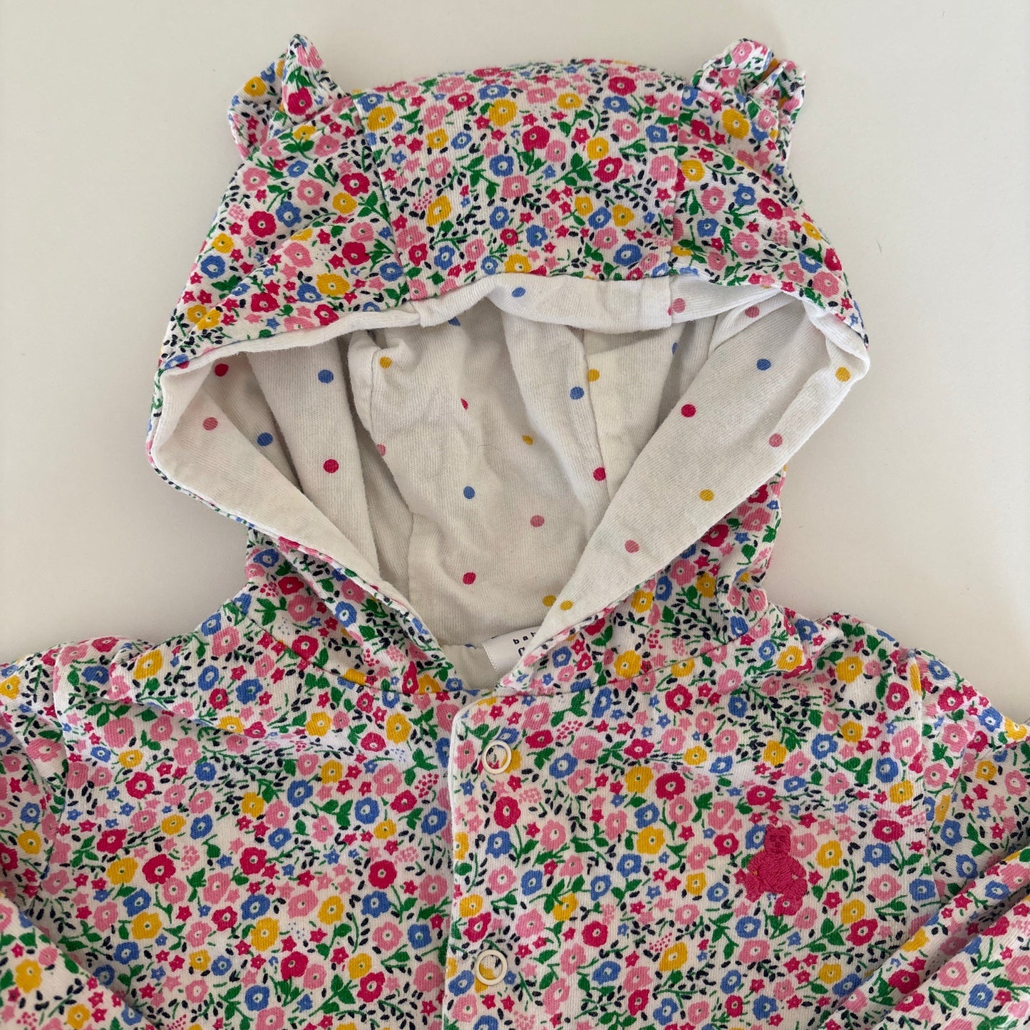 A children's hooded jacket or coat with a dense multicolor floral print on a white base, featuring bear-ear shaped hood details and a light cream inner lining with small colorful dots.