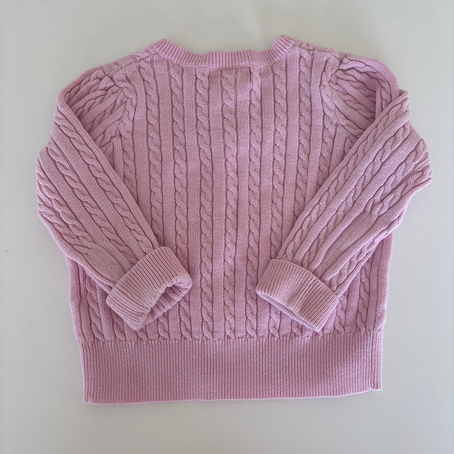 A light pink cable-knit sweater with matching ribbed cuffs and a ribbed hem, laid flat on a light off-white background.