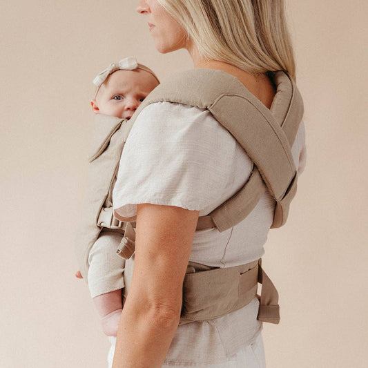 Woman carrying a baby in a beige baby carrier against a plain background