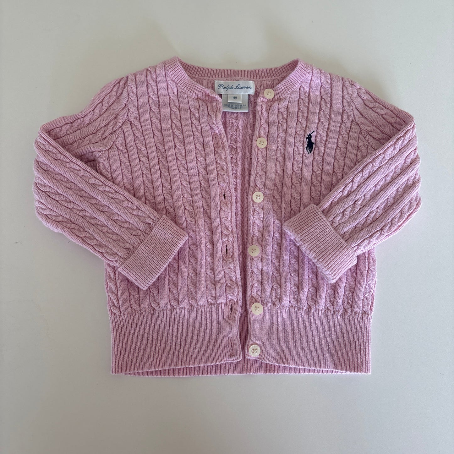 A light pink cable-knit baby cardigan sweater with a button-up front, laid flat on a light beige background. A small embroidered polo player logo is visible on the chest, identifying the brand as Ralph Lauren.