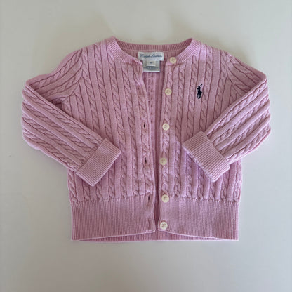 A light pink cable-knit baby cardigan sweater with a button-up front, laid flat on a light beige background. A small embroidered polo player logo is visible on the chest, identifying the brand as Ralph Lauren.
