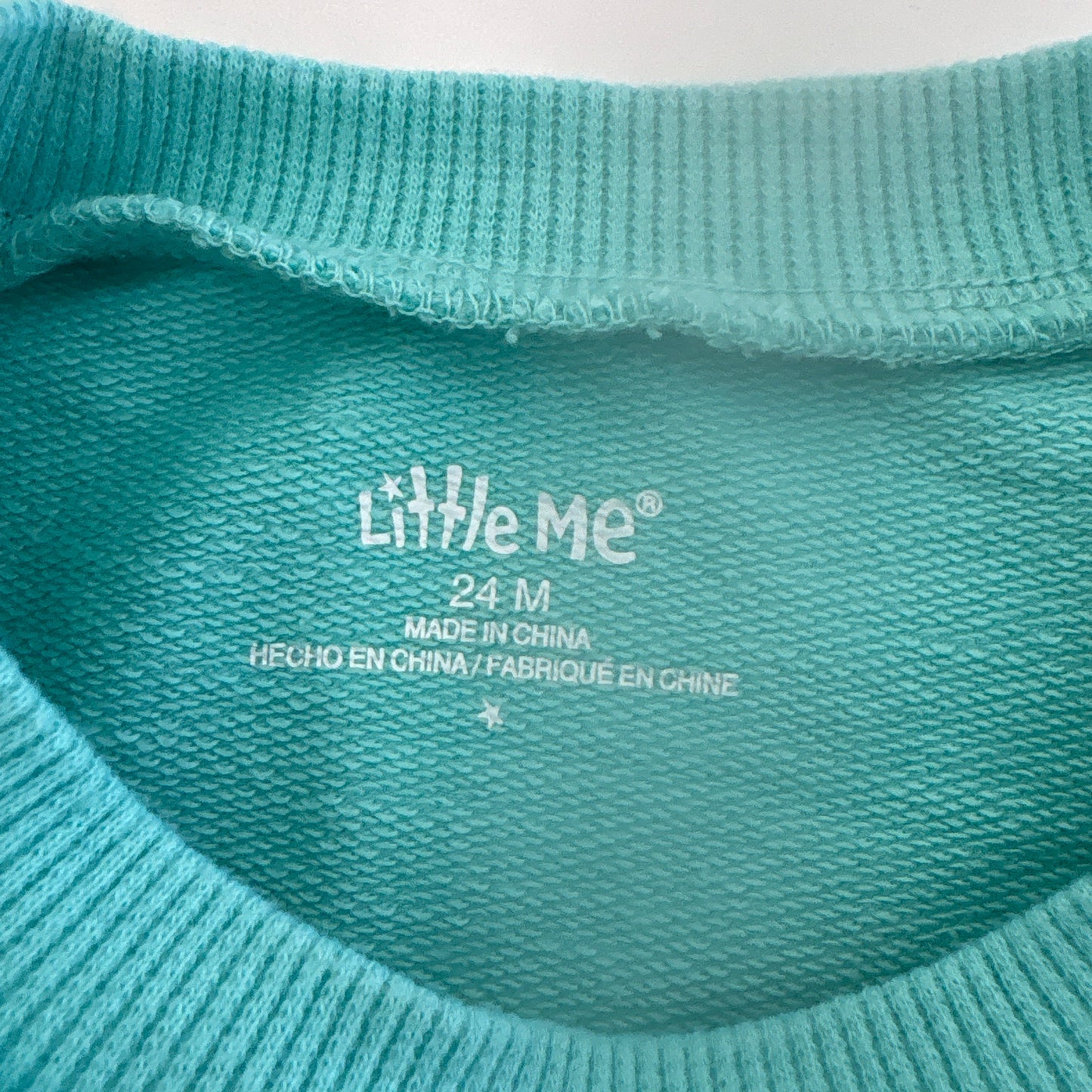 Close-up of a teal fabric with 'Little Me' branding on a light background