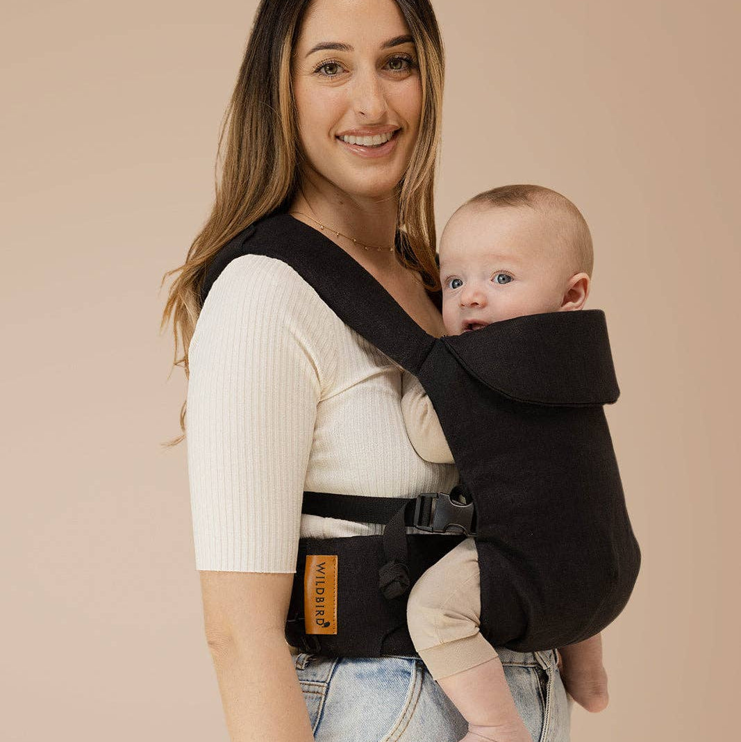Woman holding a baby in a black baby carrier against a beige background