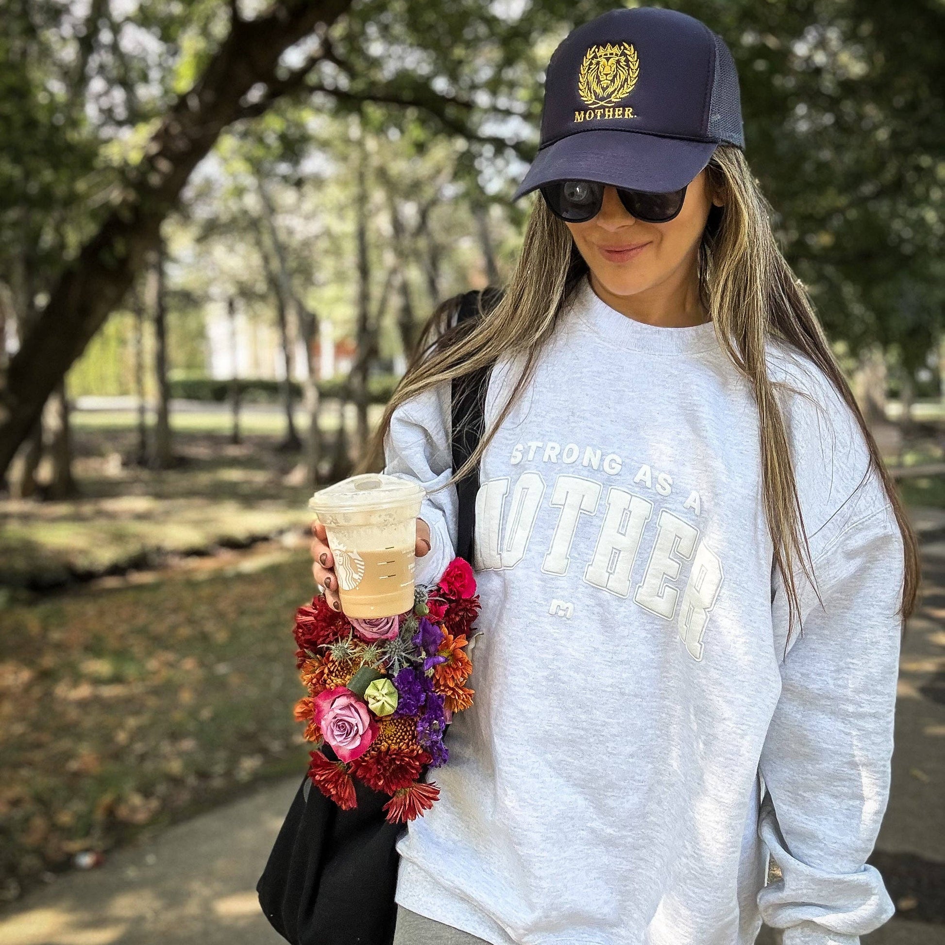 Woman in a park wearing a cap, sunglasses, and holding a drink and flowers.
