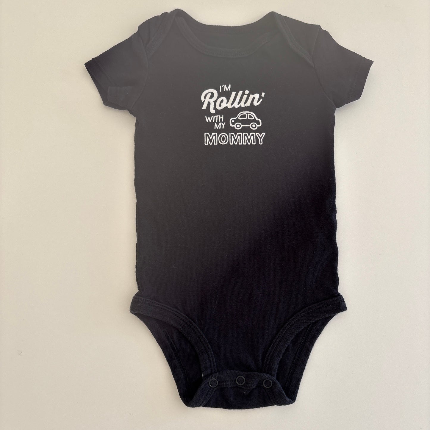 Black baby onesie with white text that says "I'm Rollin' with my Mommy" and shows a picture of a car on the onesie; on a light gray background