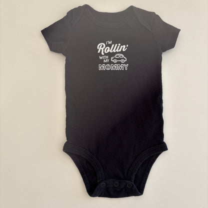 Black baby onesie with white text that says "I'm Rollin' with my Mommy" and shows a picture of a car on the onesie; on a light gray background