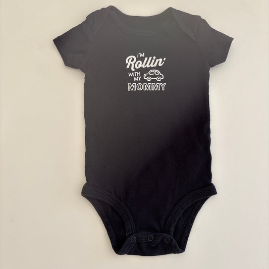 Black baby onesie with white text that says "I'm Rollin' with my Mommy" and shows a picture of a car on the onesie; on a light gray background