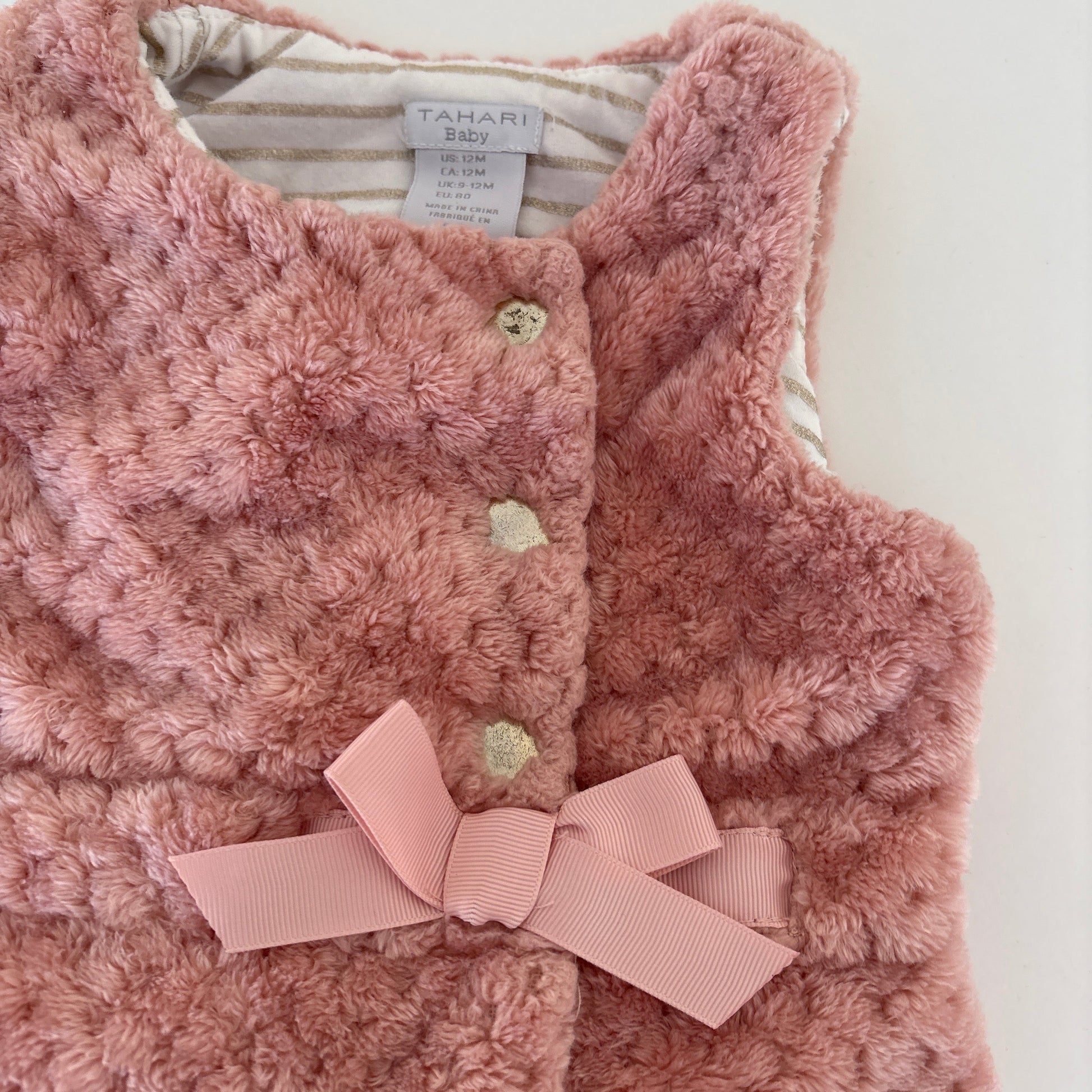 A close-up of a pink fuzzy textured vest with a matching pink ribbon bow at the center front, featuring three decorative buttons and a striped lining visible inside the collar. The brand label reads TAHARI.