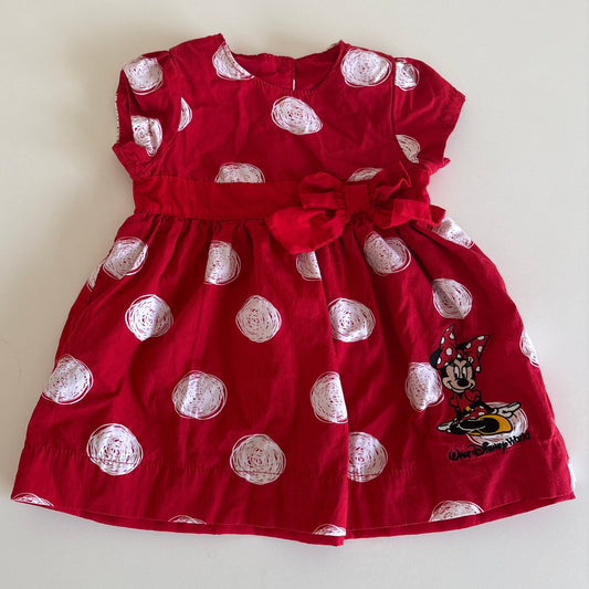 A red baby girl's dress with white polka dots, featuring a matching red waist tie bow, a Minnie Mouse graphic on the lower right, and a round neckline, laid flat on a light beige background.