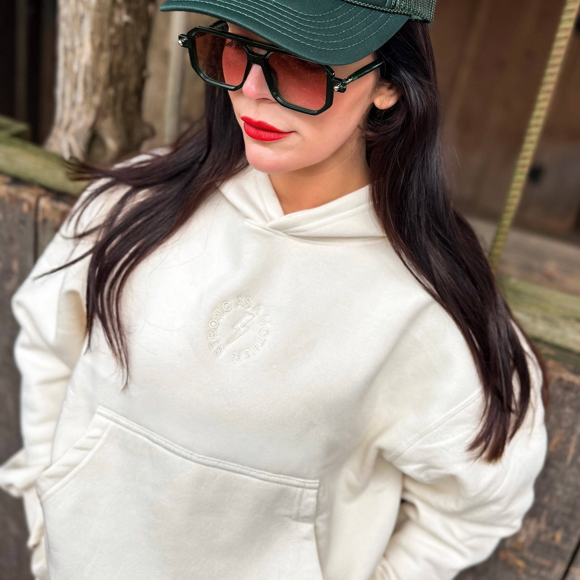 Person wearing a white hoodie, green cap, and sunglasses with a wooden background