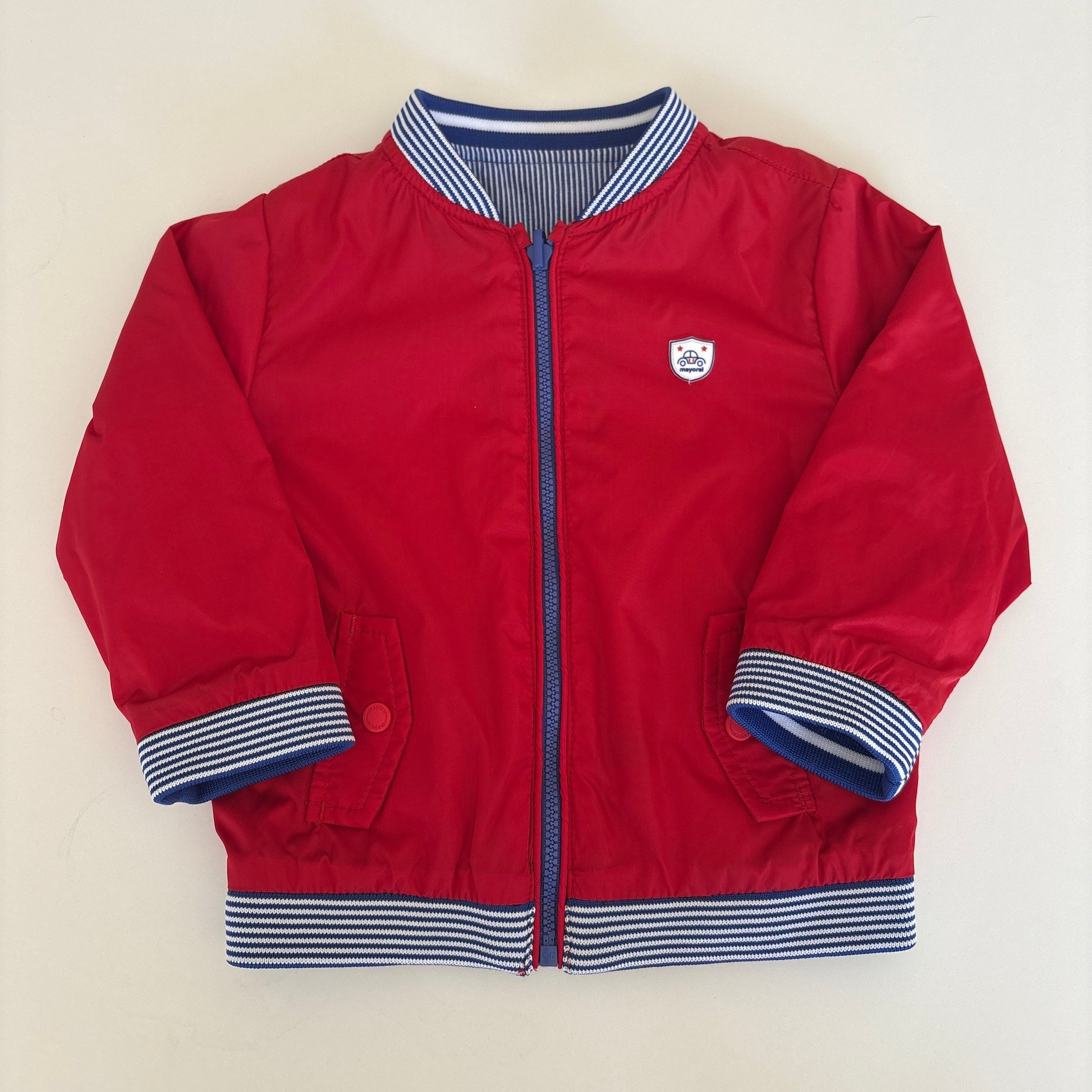 A red zip-up jacket with blue and white striped ribbed cuffs and collar, laid flat on a light beige background.