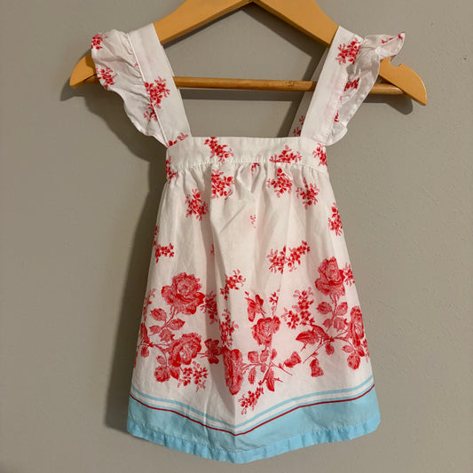 A toddler's summer dress with white, red, and blue colors, featuring floral patterns and strap details, displayed on a wooden hanger.