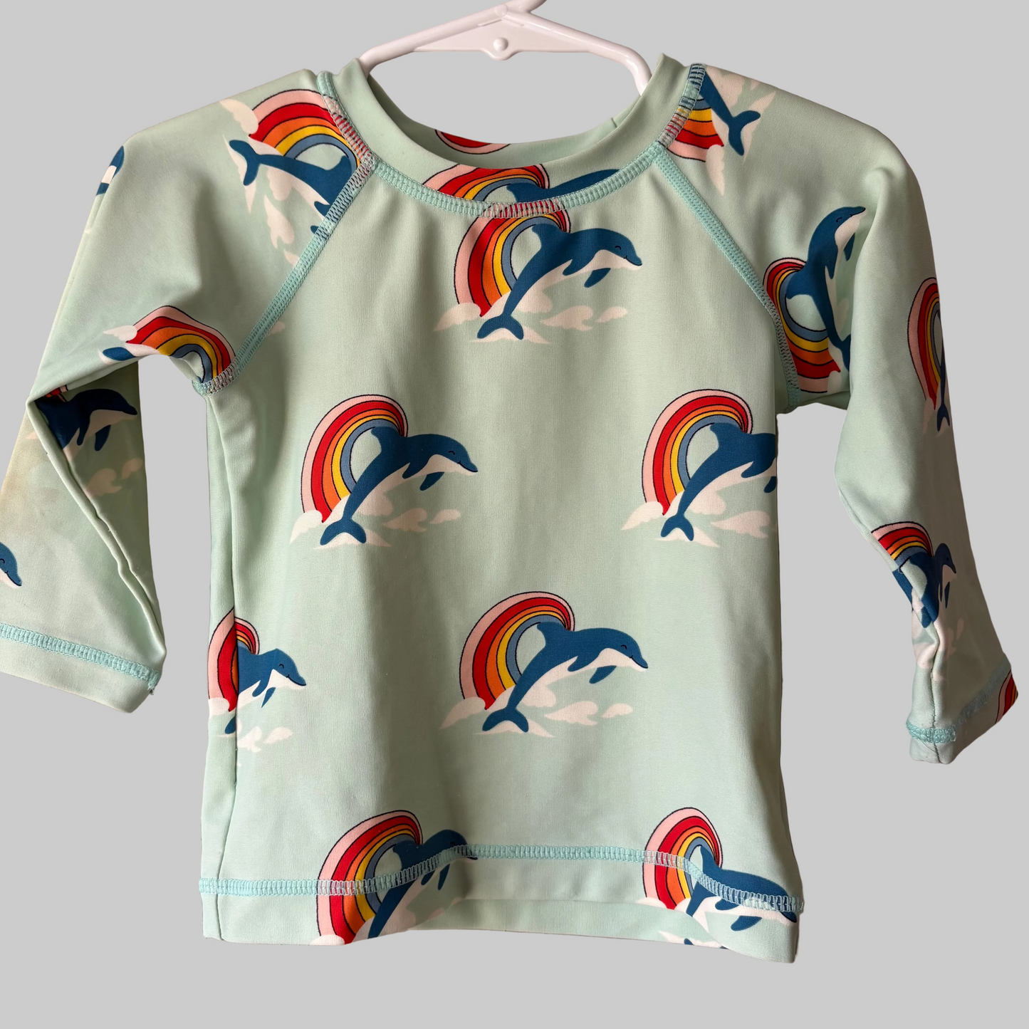 Long sleeve swim shirt for toddlers with a pattern of dolphins and rainbows on a gray background, displayed on a hanger.