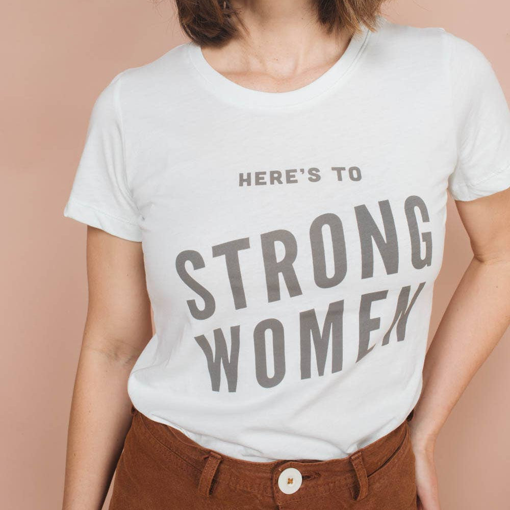 Person wearing a white t-shirt with 'Here's to Strong Women' text on a beige background