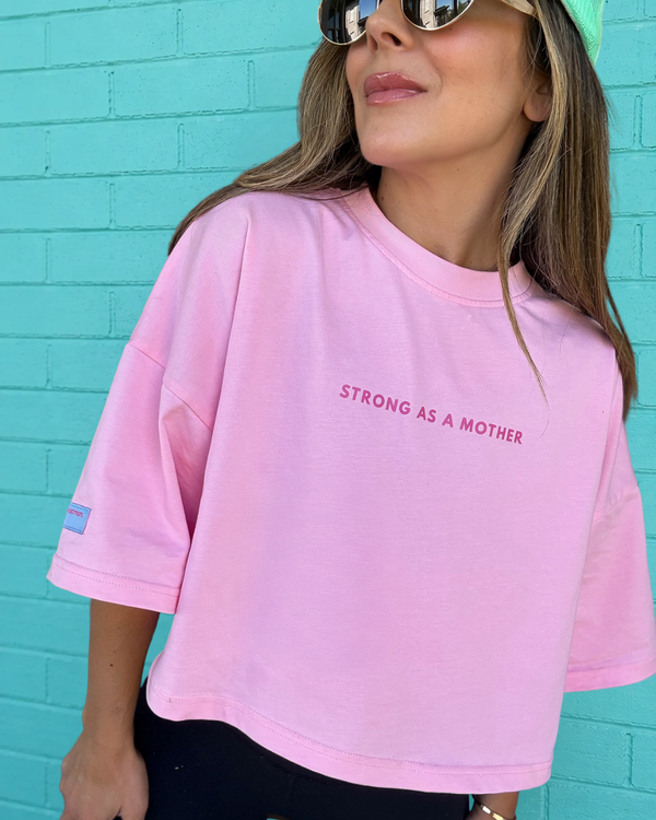 Person wearing a pink t-shirt with 'STRONG AS A MOTHER' text against a turquoise brick wall.