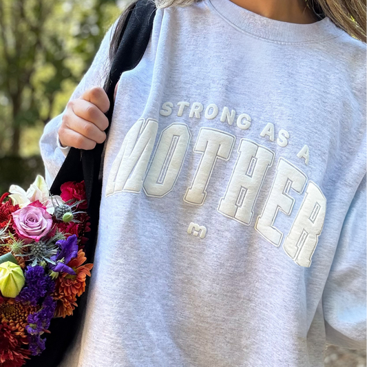 Person wearing a gray sweatshirt with 'STRONG AS A MOTHER' text, holding flowers outdoors.