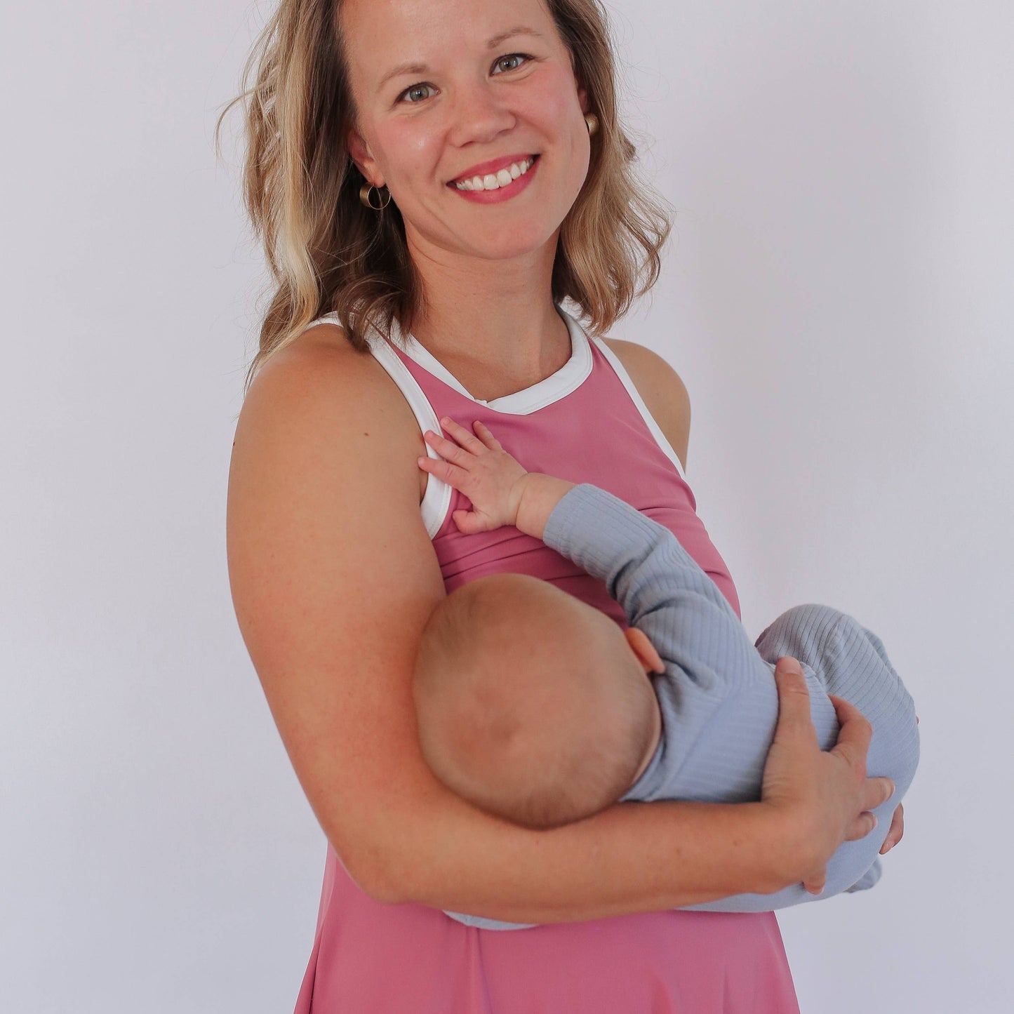 Woman in a pink nursing dress holding and breastfeeding a baby against a white background