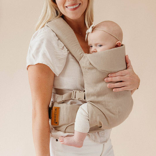 Woman holding a baby in a beige baby carrier against a plain background